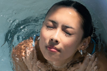 Close-up of a woman immersed in a cold plunge bath with eyes closed, water rippling around her face and shoulders in bright natural light.
