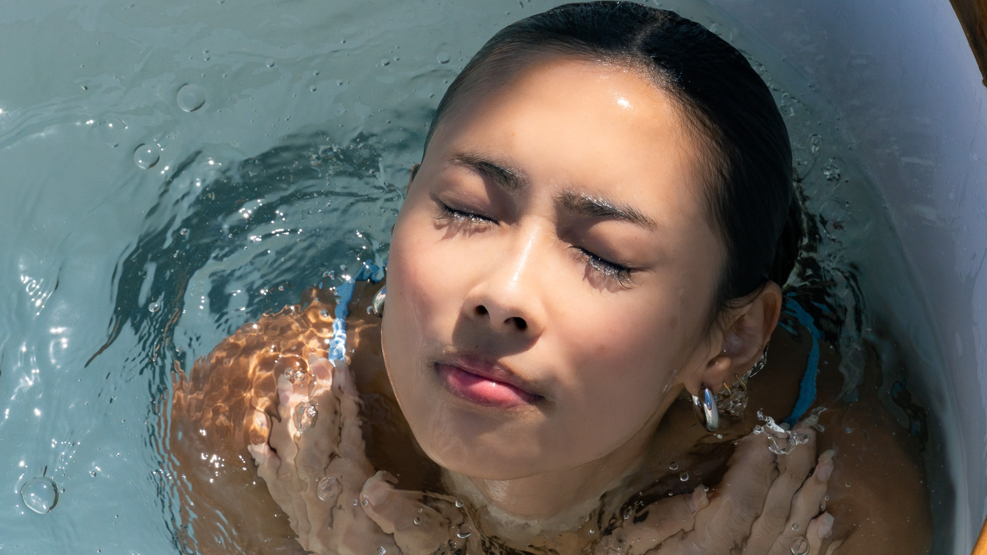 Close-up of a woman immersed in a cold plunge bath with eyes closed, water rippling around her face and shoulders in bright natural light.