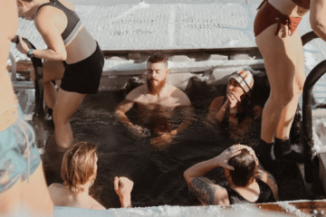 Finnish winter swimming group outside a lakeside sauna, steam rising into cold air beside the water