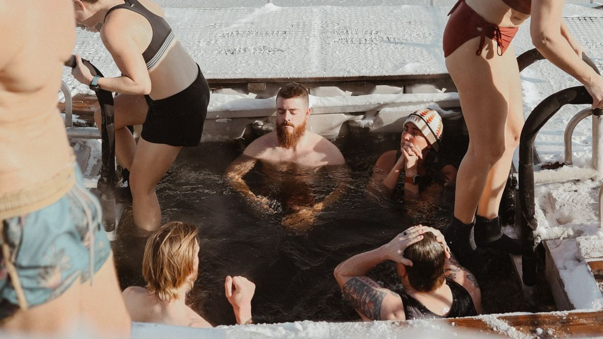 Finnish winter swimming group outside a lakeside sauna, steam rising into cold air beside the water