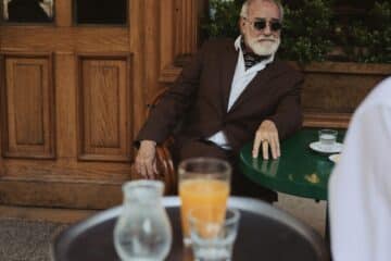 an old man sat at a table in paris, drinking orange juice.