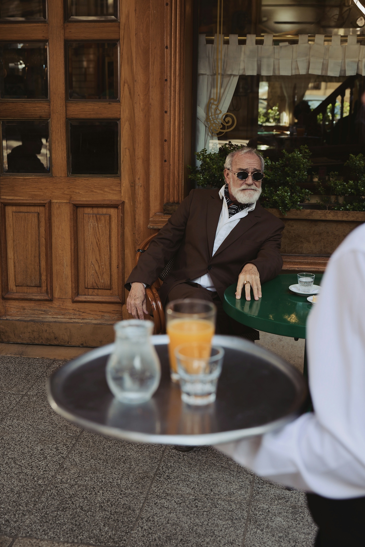 an old man sat at a table in paris, drinking orange juice.